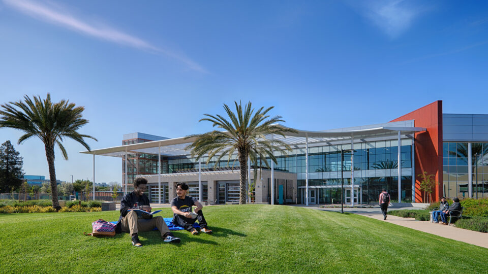 Two students sitting on a campus lawn in front of a modern glass building and palm trees under a clear blue sky