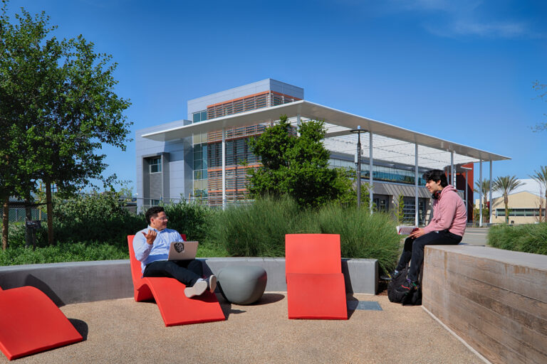 Two students sit and study in an outdoor courtyard with red chairs beside a modern campus building.