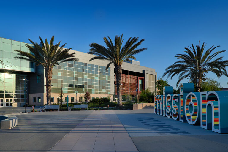 Modern glass office building with palm trees and a colorful Mission sign in an open plaza under a clear blue sky