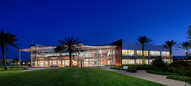 Modern glass-fronted building at dusk, surrounded by palm trees, lawn, and walkways under a deep blue sky.