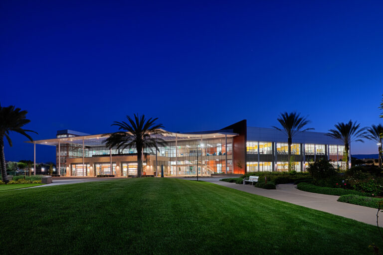 Modern glass-fronted building at dusk, surrounded by palm trees, lawn, and walkways under a deep blue sky.