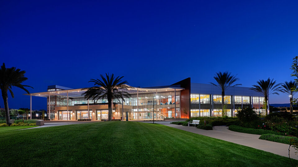 Modern glass-fronted building at dusk, surrounded by palm trees, lawn, and walkways under a deep blue sky.
