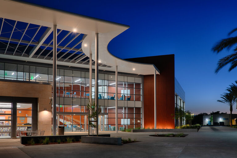 Modern glass-fronted building at dusk with a sweeping white canopy, illuminated interior, and palm-lined walkway