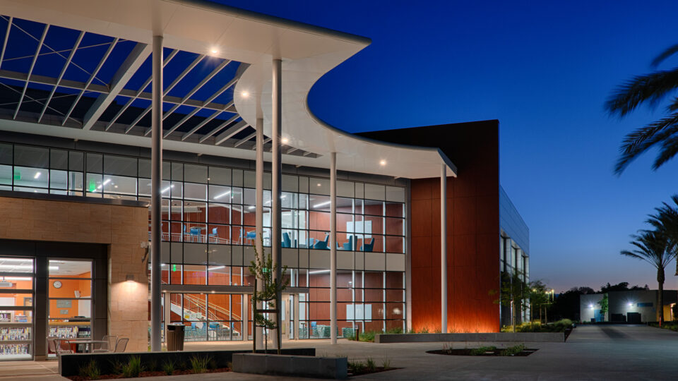 Modern glass-fronted building at dusk with a sweeping white canopy, illuminated interior, and palm-lined walkway