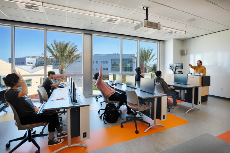 Students at computer desks raise their hands as an instructor teaches in a bright classroom with large windows and palm trees outside.