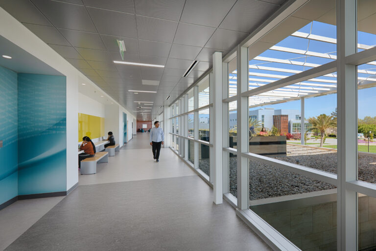 Bright modern hallway with floor-to-ceiling windows, a man walking, and students seated at built-in desks.
