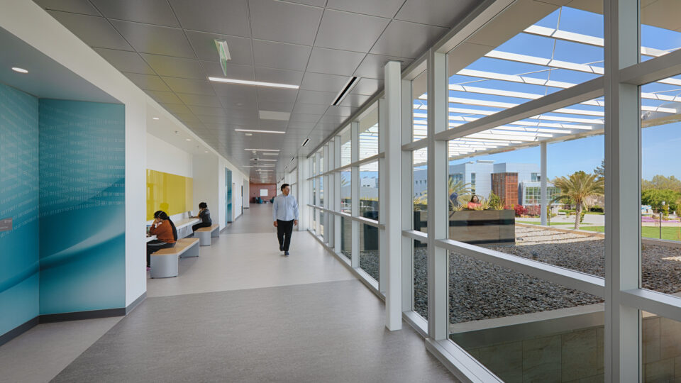 Bright modern hallway with floor-to-ceiling windows, a man walking, and students seated at built-in desks.