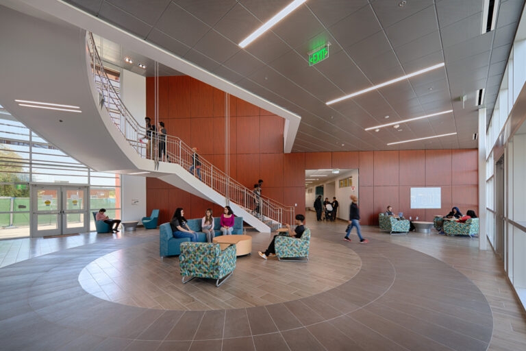 Modern building lobby with a sweeping white staircase, wood-paneled walls, and students sitting and walking around.