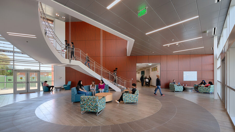 Modern building lobby with a sweeping white staircase, wood-paneled walls, and students sitting and walking around.
