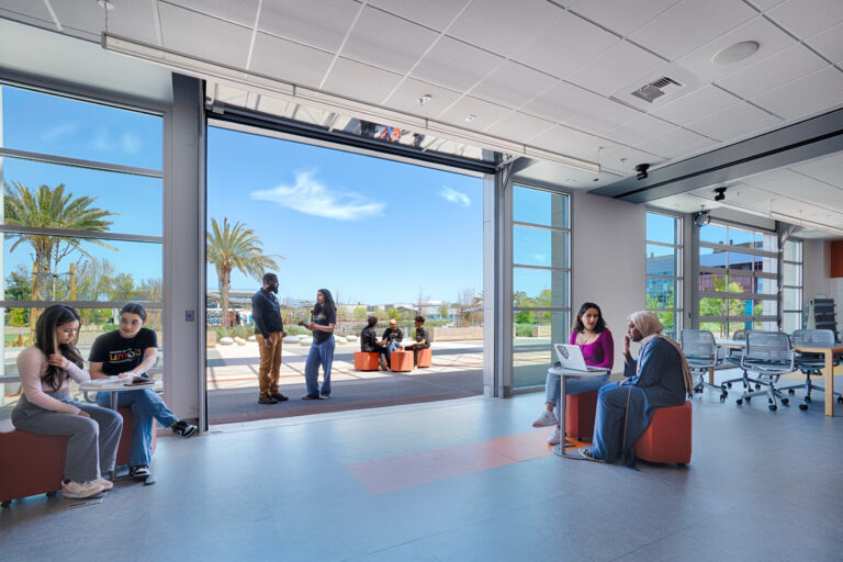 Students studying and talking in a bright indoor-outdoor campus lounge with large open doors and palm trees outside.