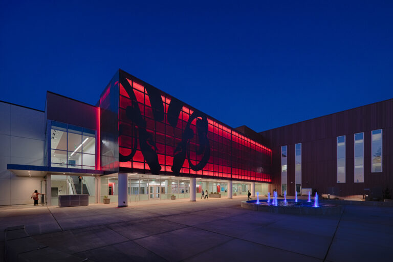 Modern building at dusk with a glowing red glass facade, open courtyard, and blue-lit fountain in front