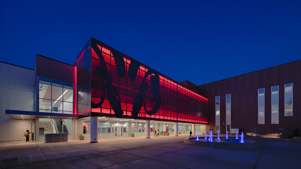 Modern building at dusk with a glowing red glass facade, open courtyard, and blue-lit fountain in front