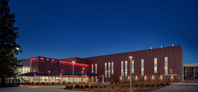 Modern campus building at dusk with a red-lit glass facade, tall narrow windows, and walkway lights in front