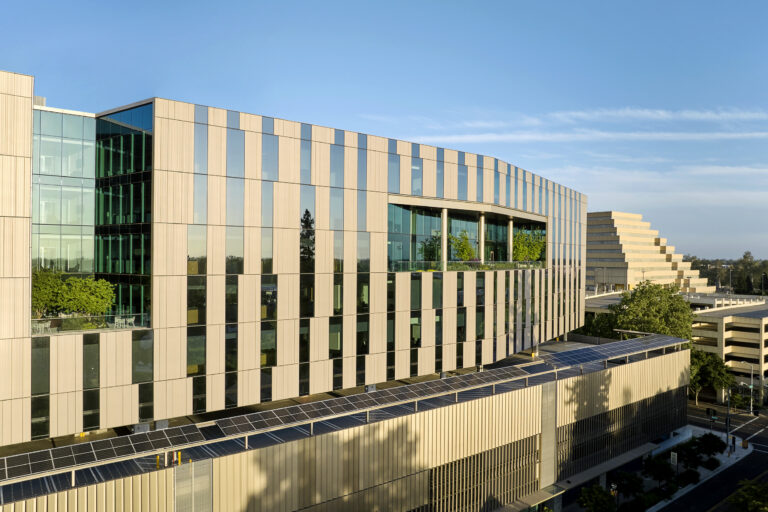 Modern office building with striped beige facade, tall glass windows, and rooftop solar panels under a clear blue sky