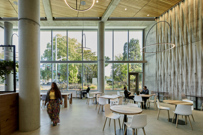 Modern café interior with tall windows, concrete columns, round tables, and a few people seated and walking.