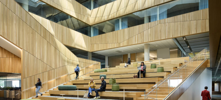 Modern wood-paneled atrium with tiered seating, skylights, and people studying and talking on the steps