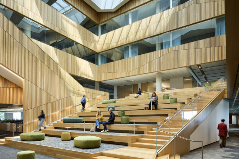 Modern wood-paneled atrium with tiered seating, skylights, and people studying and talking on the steps