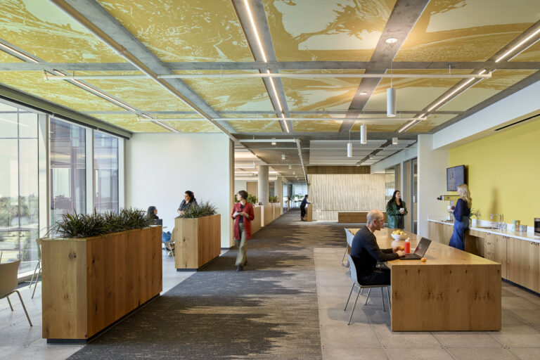 Modern open office lounge with wooden planters and tables, large windows, and people working, walking, and making coffee