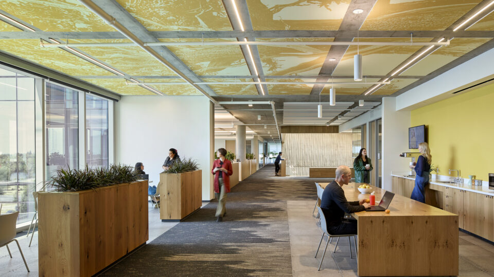 Modern open office lounge with wooden planters and tables, large windows, and people working, walking, and making coffee