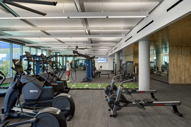 Modern gym interior with cardio machines, rowing machines, weight equipment, and two people exercising by large windows