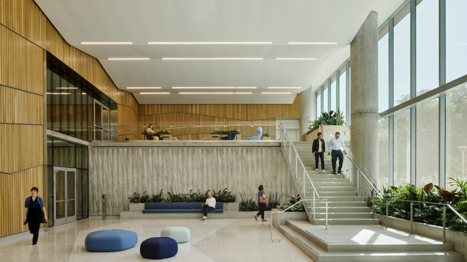 Bright modern atrium with wood-paneled walls, tall windows, concrete stairs, plants, and people walking or sitting.