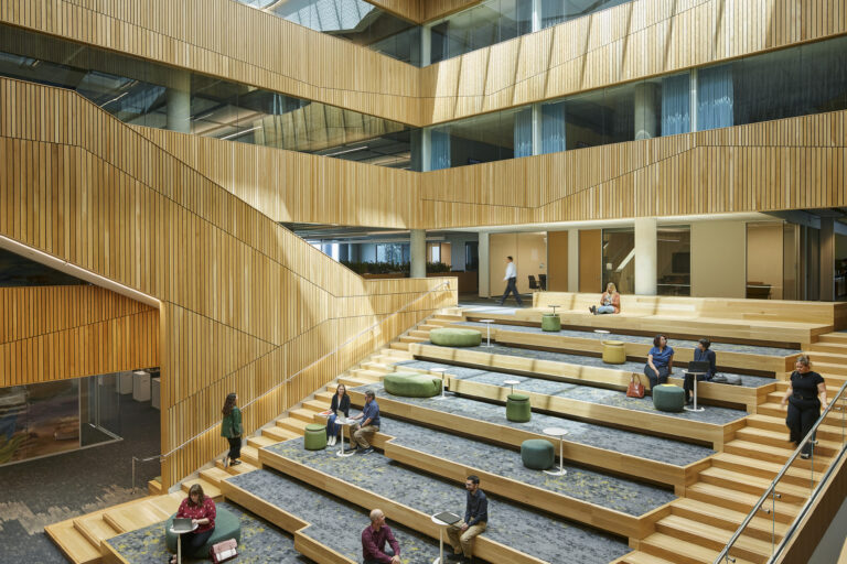 Spacious modern atrium with tiered wooden seating, skylights, and people sitting, talking, and working on laptops