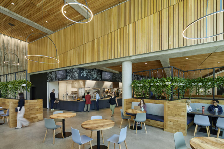 Modern cafeteria interior with wood-paneled walls, circular pendant lights, plant-lined booths, and people dining and ordering food.