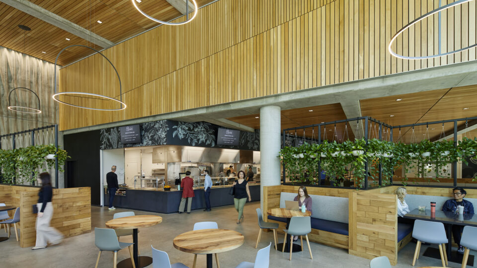 Modern cafeteria interior with wood-paneled walls, circular pendant lights, plant-lined booths, and people dining and ordering food.