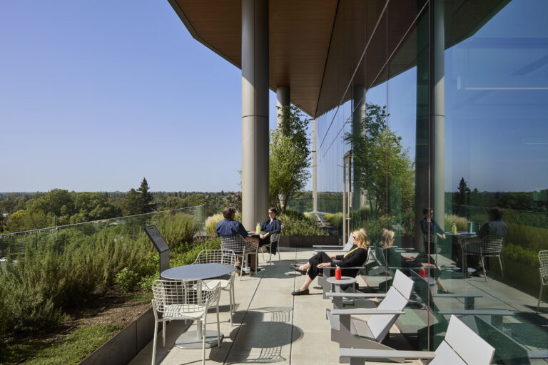 People relaxing on a modern glass-walled terrace with chairs, tables, and views of trees under a clear blue sky