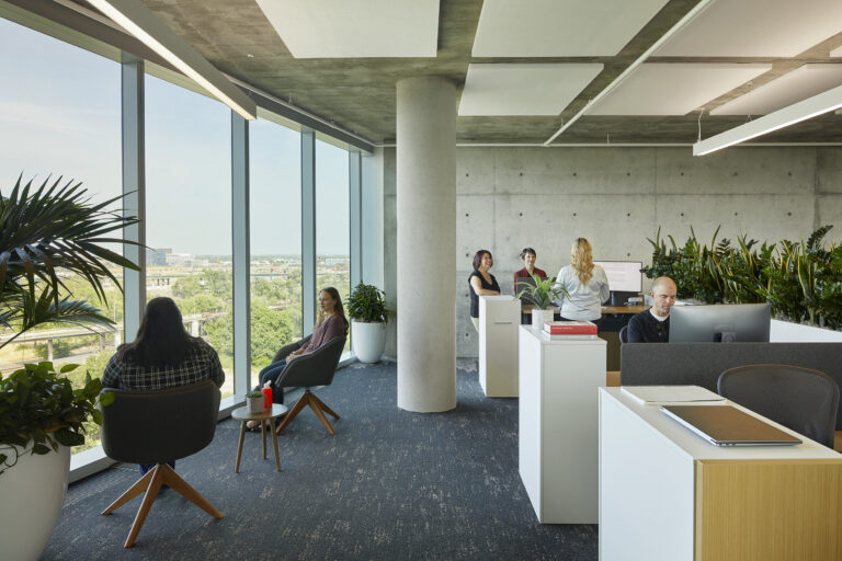Modern open-plan office with large windows, plants, and employees sitting, talking, and working at desks