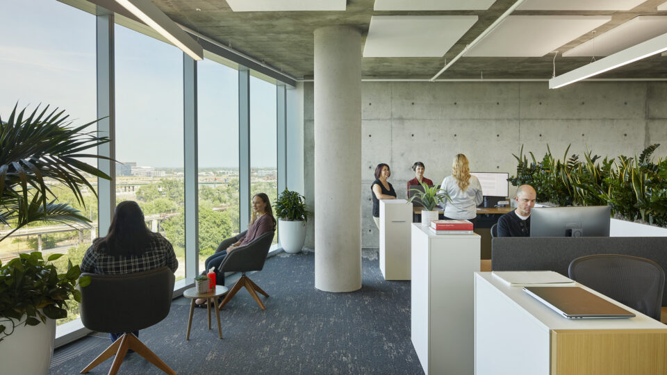 Modern open-plan office with large windows, plants, and employees sitting, talking, and working at desks