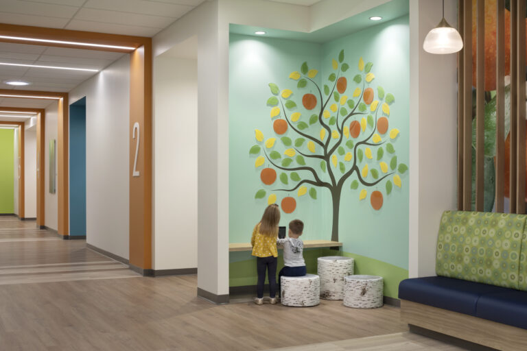 Two children stand at a small desk beneath a colorful fruit tree mural in a bright hallway.
