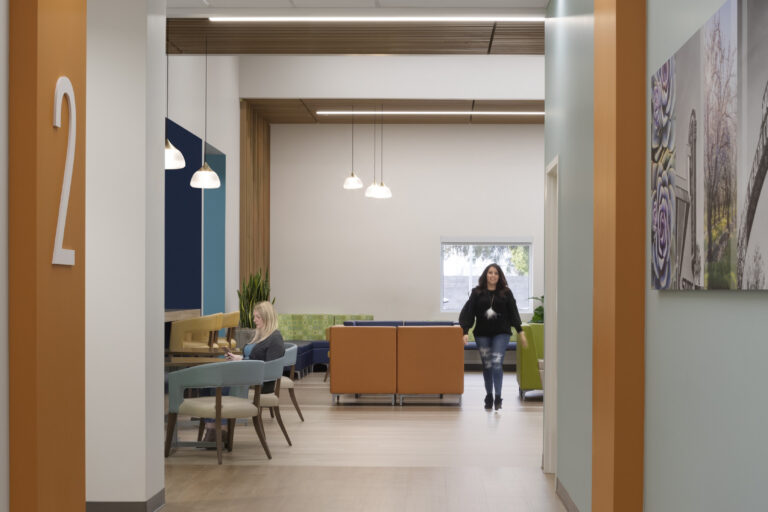 Modern waiting area viewed through an orange-framed doorway marked 2, with colorful seating and two women inside