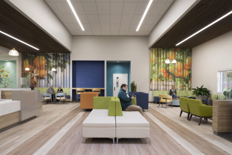 Bright modern waiting room with colorful seating, wood floors, and orange tree wall murals, with several people seated inside