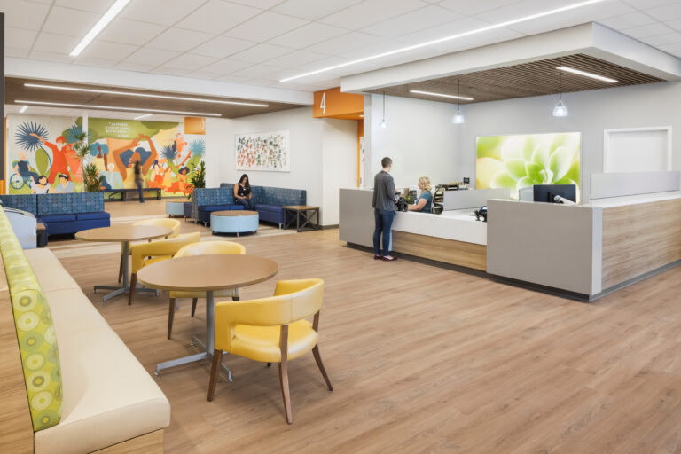 Bright modern reception and waiting area with seating, wood floors, colorful mural, and a person checking in at the desk
