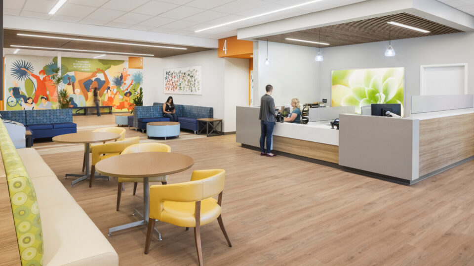 Bright modern reception and waiting area with seating, wood floors, colorful mural, and a person checking in at the desk