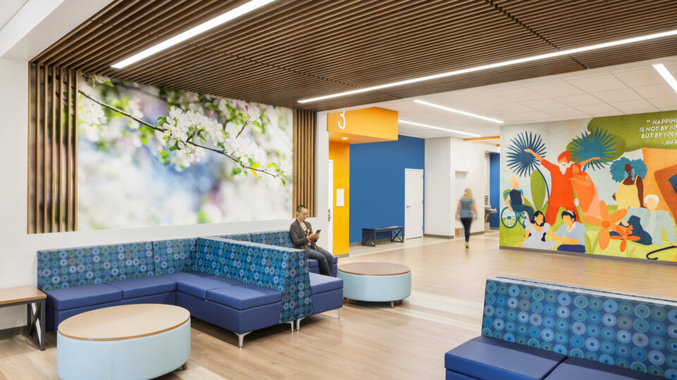 Modern waiting area with blue booth seating, wood-slat ceiling, floral wall art, and a colorful mural on the right.