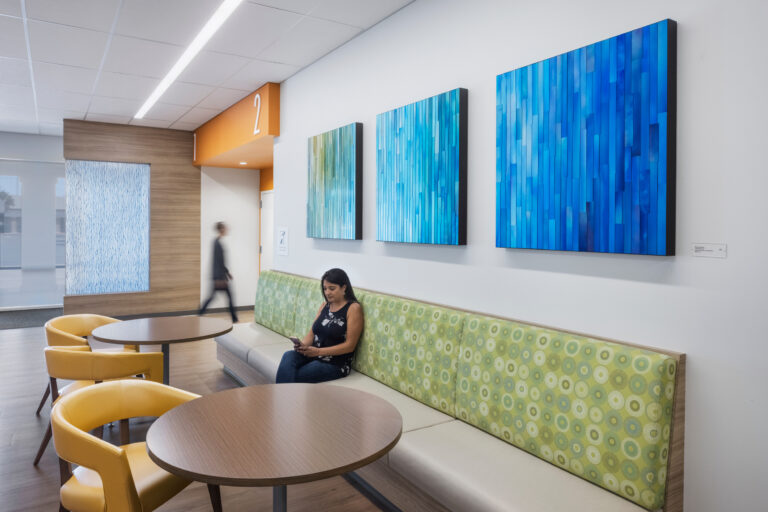 Modern waiting area with round tables, yellow chairs, a woman seated on a green bench, and blue abstract wall art