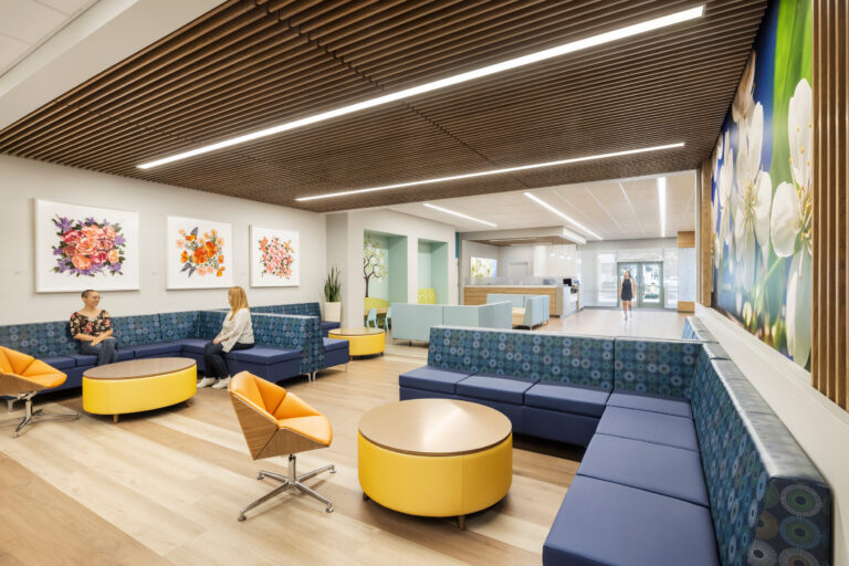 Bright modern lobby with blue booth seating, yellow tables, floral artwork, and two women talking near the reception area