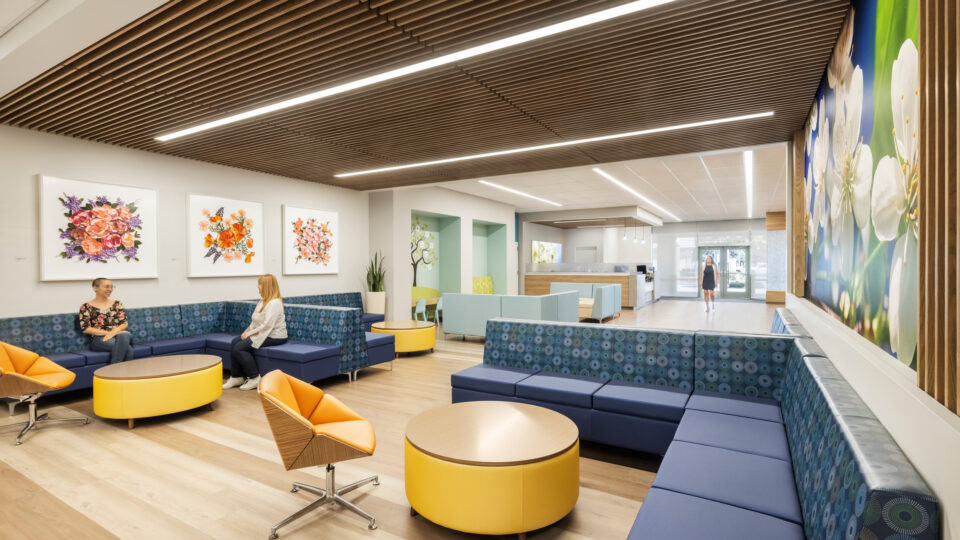 Bright modern lobby with blue booth seating, yellow tables, floral artwork, and two women talking near the reception area