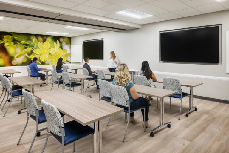 Instructor speaking to a small class in a modern training room with tables, chairs, large screens, and a flower mural.