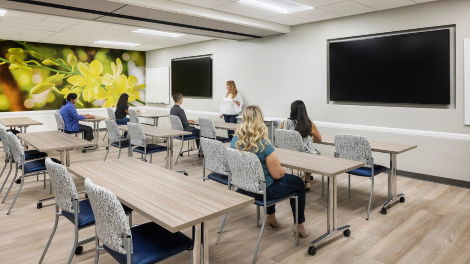 Instructor speaking to a small class in a modern training room with tables, chairs, large screens, and a flower mural.