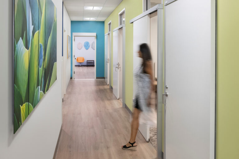 Bright office hallway with colorful walls, framed leaf artwork, and a blurred woman entering a room.