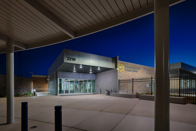 Modern Department of Corrections building entrance at dusk, with glass doors, address 2210, and a black security fence