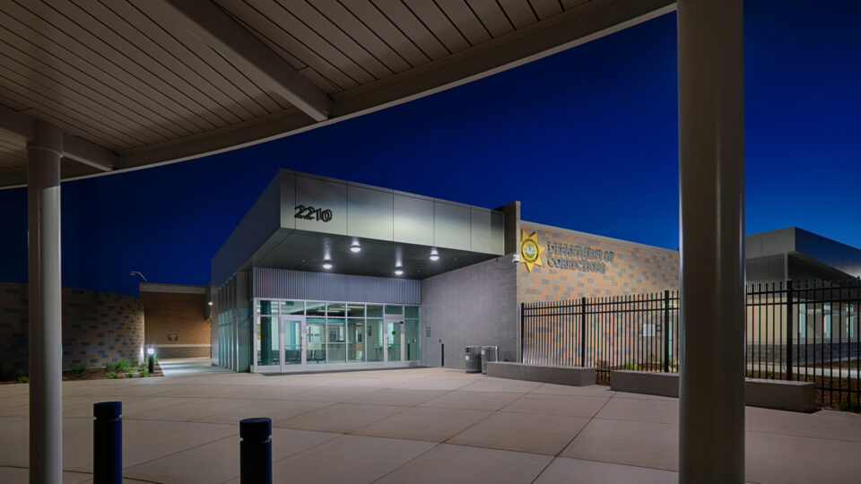 Modern Department of Corrections building entrance at dusk, with glass doors, address 2210, and a black security fence