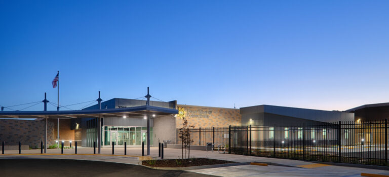 Department of Corrections building entrance at dusk, with a fenced perimeter, flagpole, and empty parking area