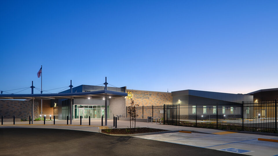 Department of Corrections building entrance at dusk, with a fenced perimeter, flagpole, and empty parking area