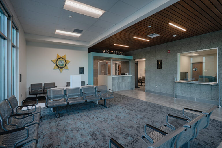 Modern visitation waiting area with metal seating, a glass booth, and a sheriff’s star emblem on the wall.