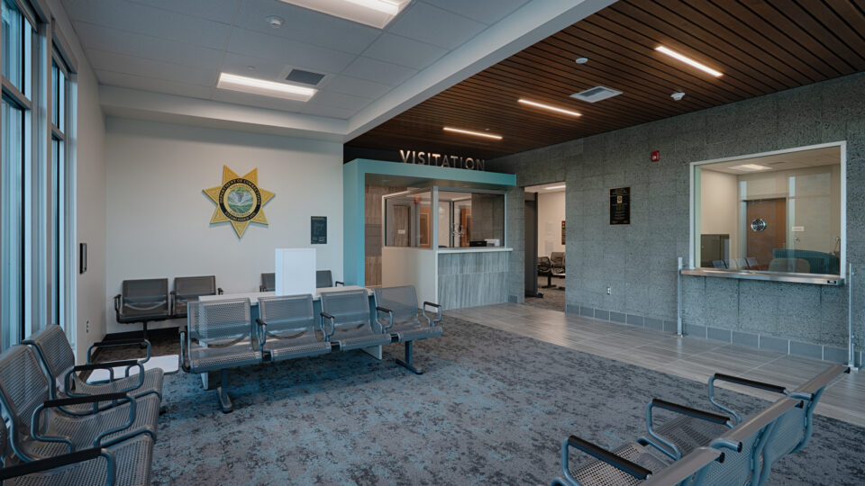 Modern visitation waiting area with metal seating, a glass booth, and a sheriff’s star emblem on the wall.