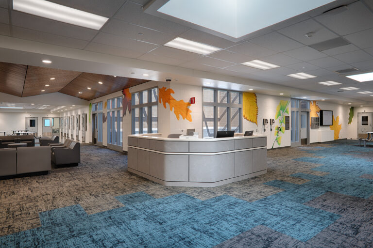 Modern reception area with curved desk, seating, blue patterned carpet, and colorful leaf murals on the walls